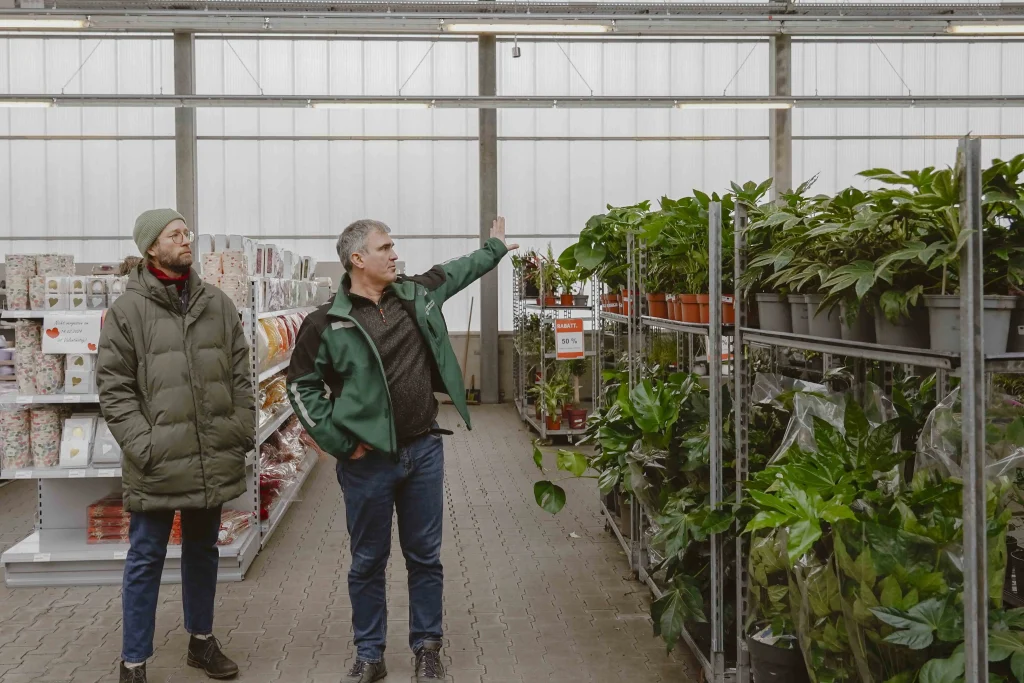 Zeigt zwei Personen im Gartenmarkt. Einer der Männer zeigt auf die Pflanzenständer.